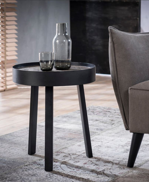 Beistelltisch TECA 45 mit runder Tischplatte in Greywashed-Optik und schwarzen Beinen in Gesamtansicht neben einem Sessel.
