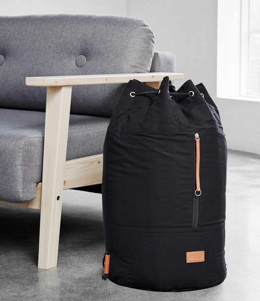 Design Wäschesack ROADIE Black in schwarzer Ausführung mit Kordelzug und Rucksacktragegurten neben grauem Sessel.
