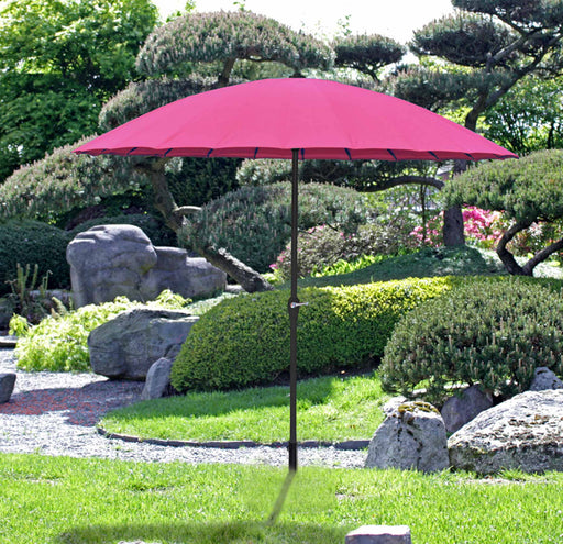 Sonnenschirm CABRERA Pink Ø 255 cm 24 Streben mit Handkurbel in Pink, aufgespannt in einem Garten mit Steinen und Begrünung.
