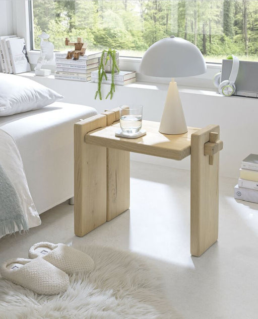 Nachtkommode Inger Eiche massiv in Gesamtansicht neben einem Bett mit Dekoobjekten auf der Ablage.