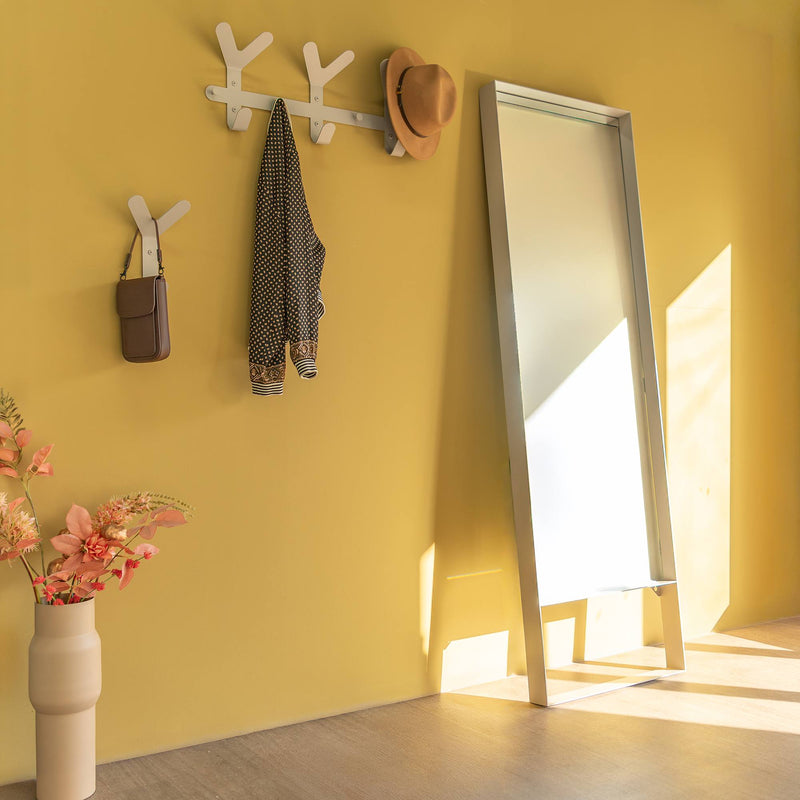 Gelbe Wand mit weißen Haken, an denen eine Tasche, ein Hemd und ein Hut hängen, daneben ein großer Spiegel