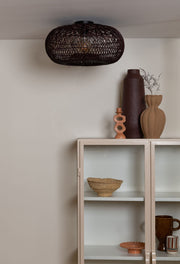 Gesamtansicht der weinroten Deckenleuchte Arizona aus Rattan mit ovalem Geflechtschirm über einem Sideboard mit Vasen.