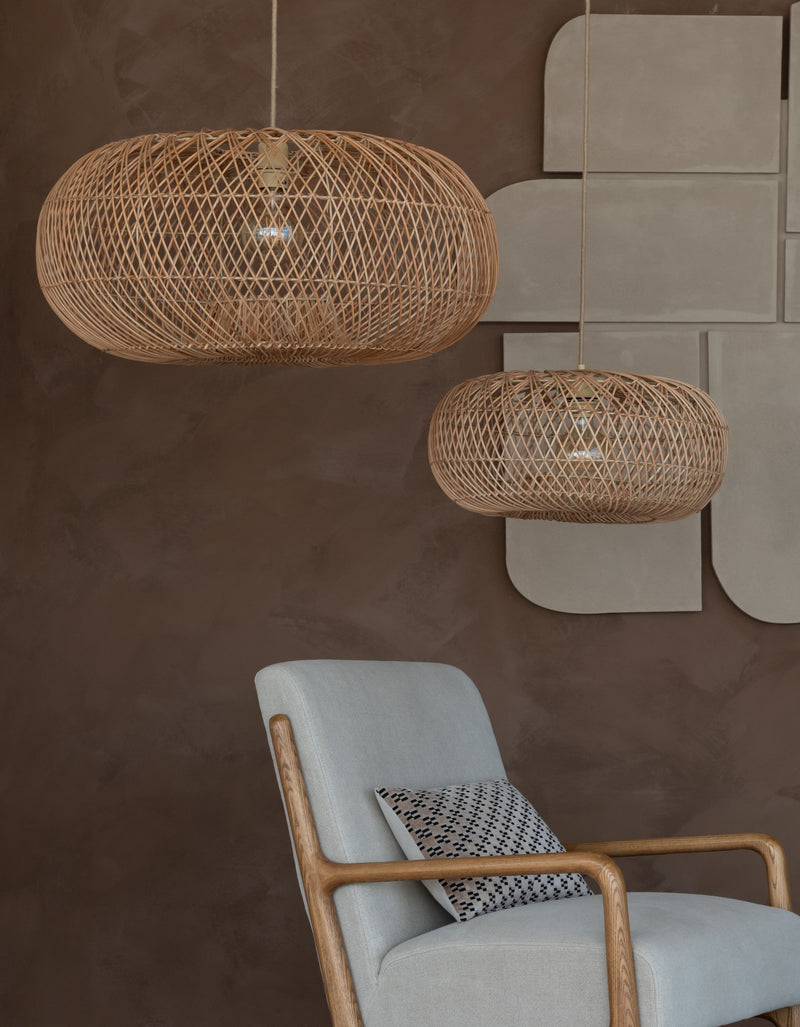 Zwei ovale Hängeleuchten "Arizona" aus geflochtenem Rattan in Naturfarbe über einem grauen Sessel mit Holzgestell.