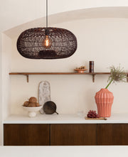 Dunkelrote Rattan-Hängeleuchte Arizona mit ovalem Geflecht, über einem Sideboard mit Holzfronten und Regalbrett hängend.