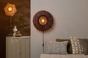 Runde Wandleuchte Arizona mit dunkel geflochtenem Rattan-Schirm, im Vordergrund leuchtend an einer hellen Wand über dem Sofa