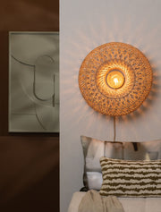 Runde Wandleuchte Arizona aus Rattan in Naturfarbe, mit eingeschalteter Glühbirne vor heller Wand, Ø 50 cm
