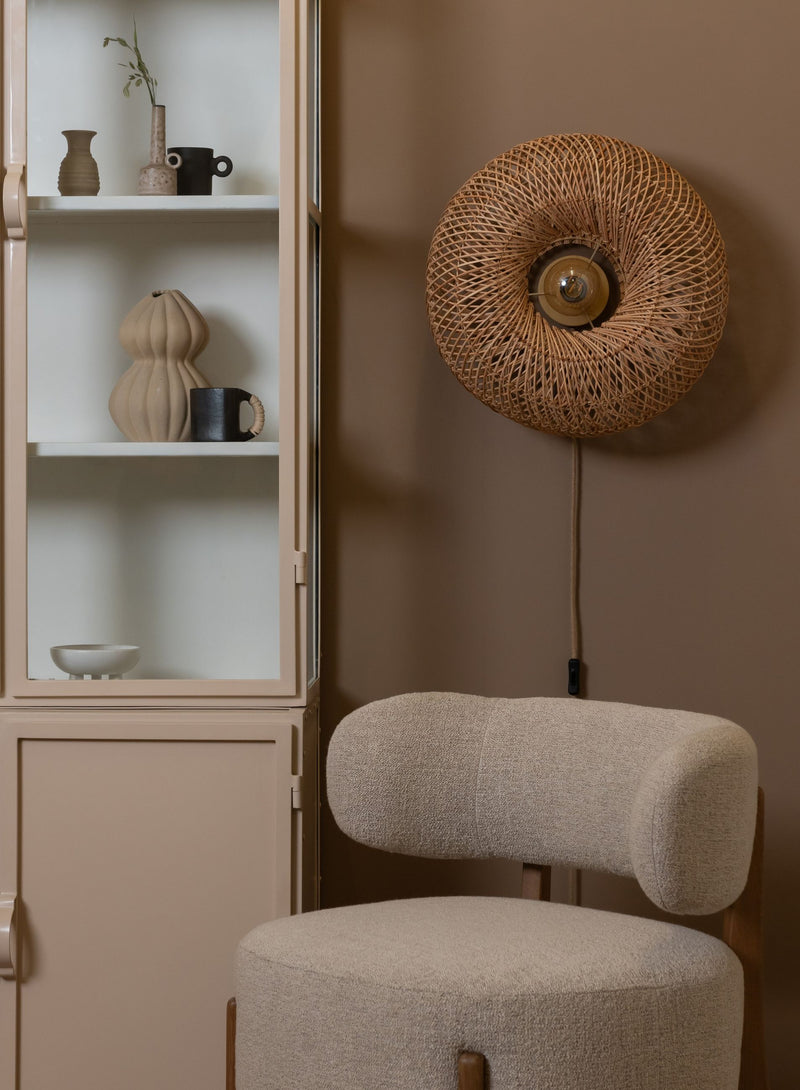 Good&Mojo Wandleuchte Arizona aus Rattan mit rundem, geflochtenem Schirm, an einer beigen Wand über einem Polsterstuhl.