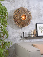 Runde Wandleuchte Bali aus Bambus in Natur/Schwarz, im Wohnraum über einem grauen Sofa montiert, mit sichtbarem Leuchtmittel.