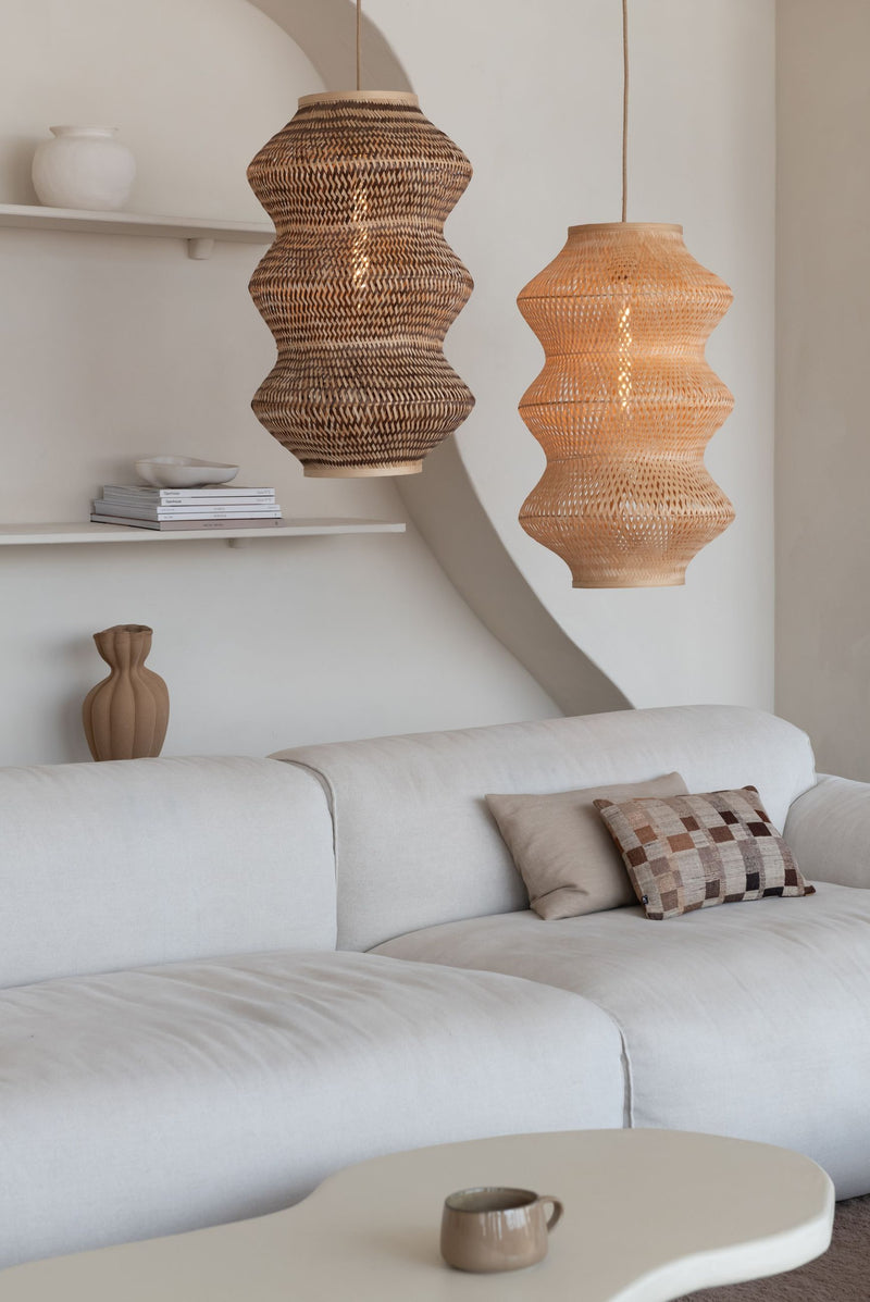 Zwei geschwungene Hängeleuchten Barbados XL aus geflochtenem Bambus in Naturtönen über einem hellen Sofa im Wohnraum.