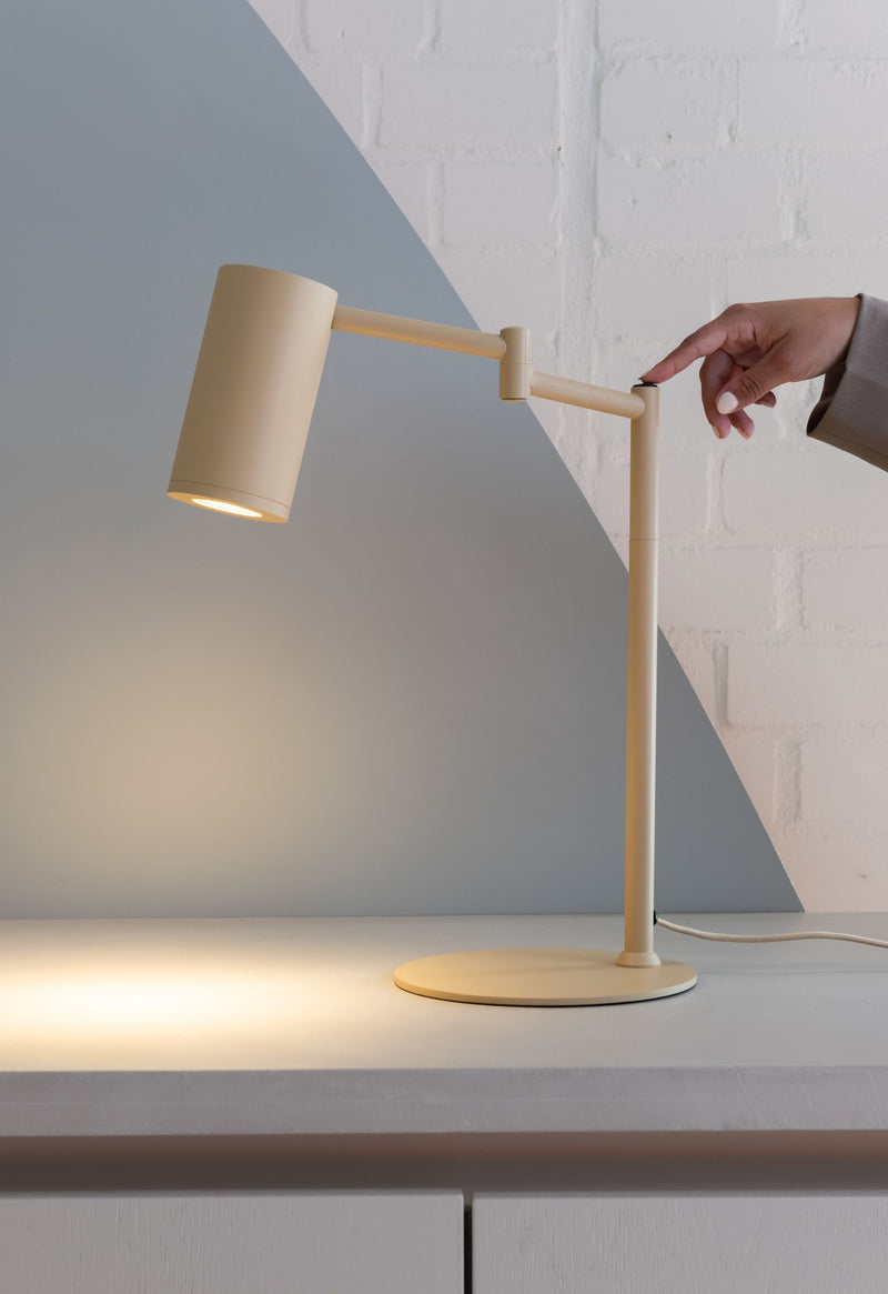 Eine beige Tischleuchte aus Eisen mit verstellbarem Arm, eingeschaltet, auf hellem Sideboard vor grauer Wand.