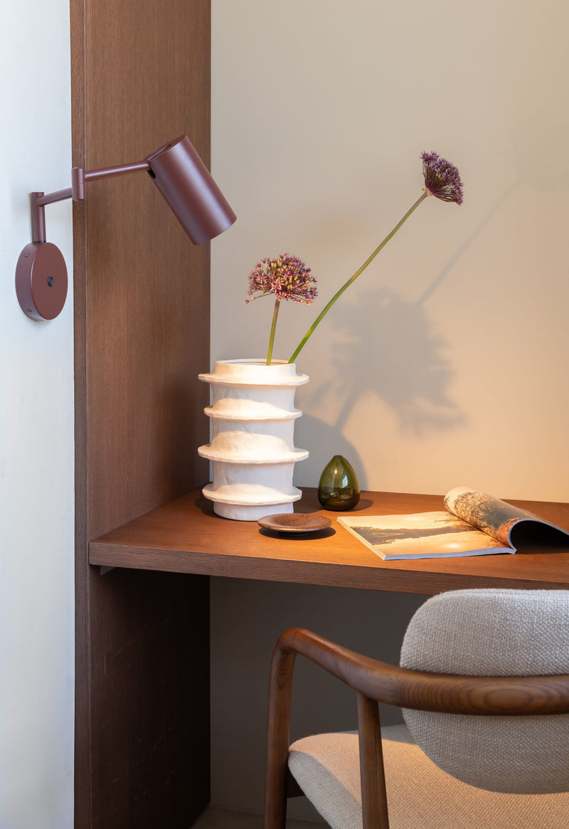 Bordeauxrote Wandleuchte "It's about RoMi Bilbao" aus Eisen mit verstellbarem Arm, über einem Schreibtisch mit Vase.