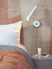 It’s about RoMi Wandleuchte Bordeaux in Weiß, mit schwenkbarem Leuchtarm, montiert über einem Bett mit Kissen und Tagesdecke.