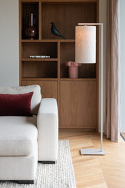 Stehleuchte It's about RoMi Boston mit hellem Leinenschirm und Eisengestell, neben einem Sofa vor einem Holzregal.