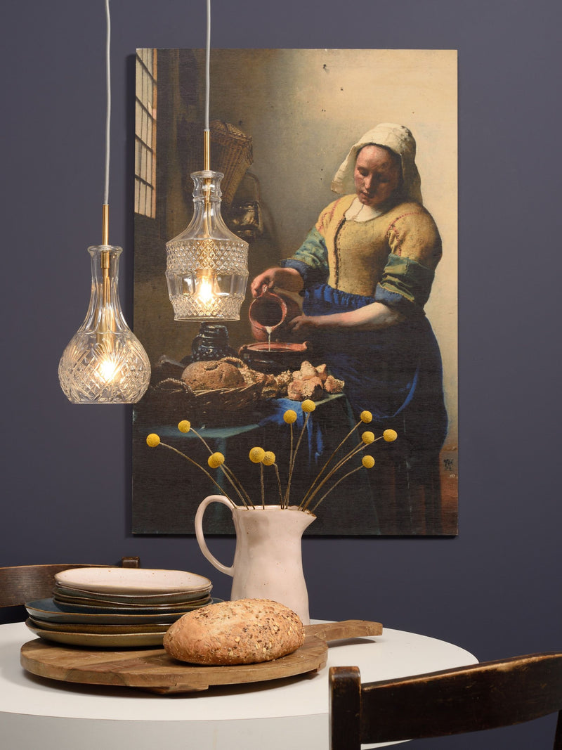 Zwei Glas-Hängeleuchten mit goldenen Fassungen über einem Esstisch, im Hintergrund ein Gemälde mit einer Frau am Krug.