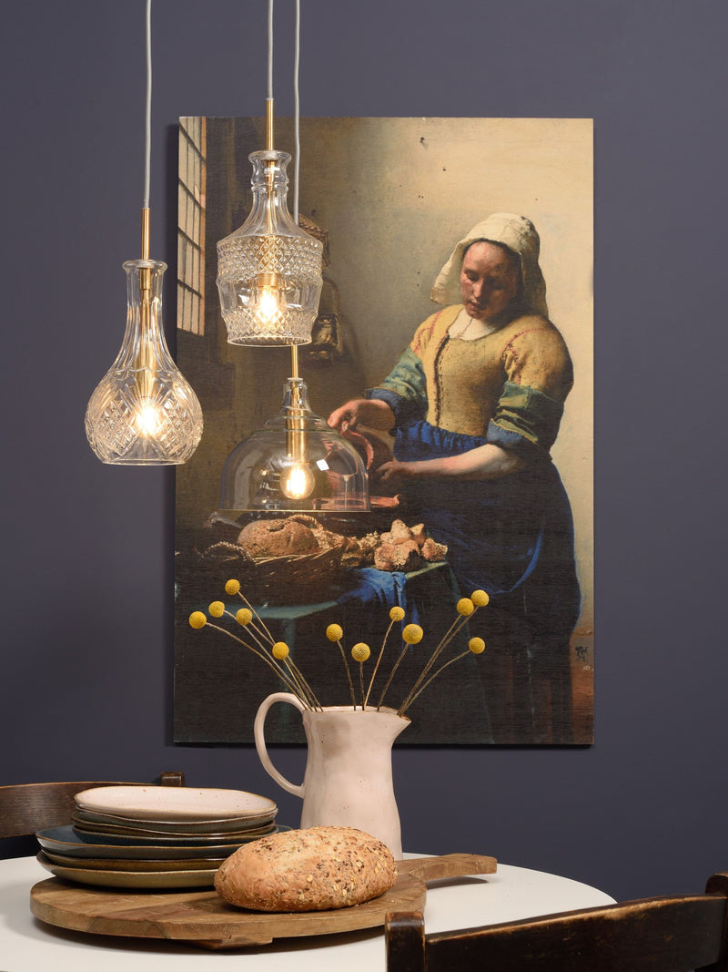 Drei Glas-Hängeleuchten mit transparentem Schliff und goldenen Details hängen vor einer Wandbild-Reproduktion mit Dienstmagd-Motiv.