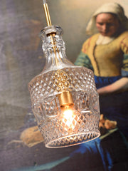 RoMi Hängeleuchte Brussels mit goldenem Fassungsteil und transparentem Glas, eingeschaltet vor einem Gemälde.