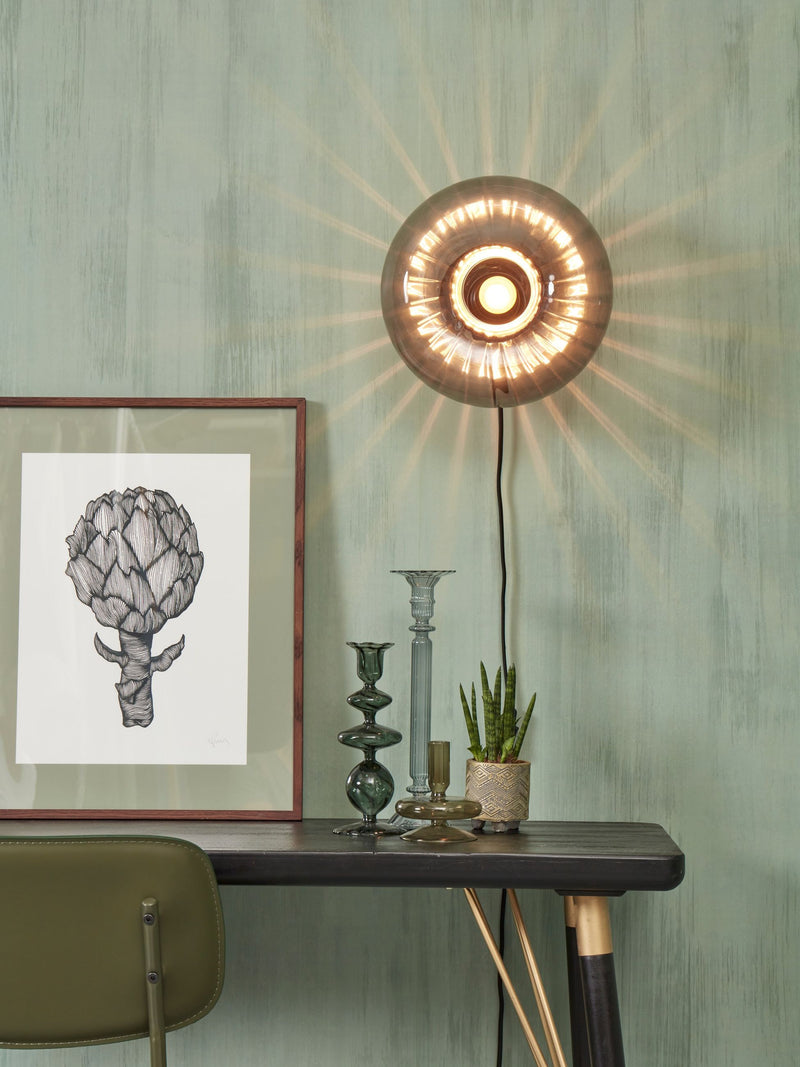 Wandleuchte It's about RoMi Brussels mit rundem, rauchgrauem Glasschirm und leuchtendem Lichtspiel an einer grünen Wand.