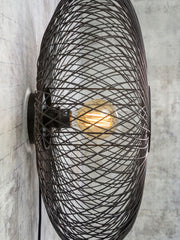 Wandleuchte Cango von Good&Mojo mit schwarzem Drahtgeflecht-Schirm und sichtbarer Glühlampe vor grauer Betonwand.