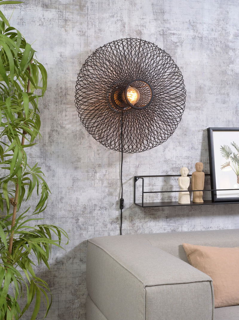 Gesamtansicht der schwarzen Wandleuchte Good&Mojo Cango Bambus Ø 60 cm mit eingeschaltetem Leuchtmittel vor grauer Betonwand.
