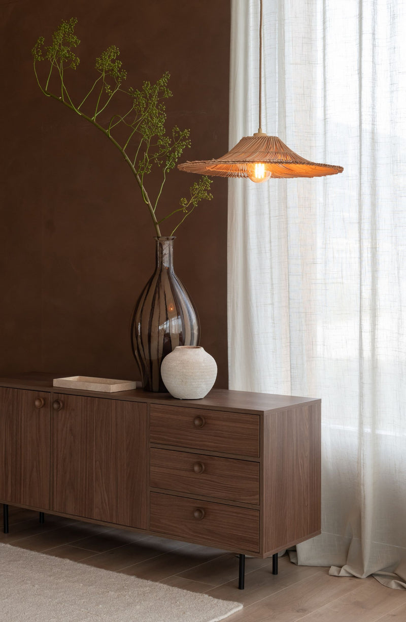 Hängeleuchte Cancun aus Naturrattan mit breitem, geflochtenem Schirm über einem Sideboard mit Glasvase und Zweig.