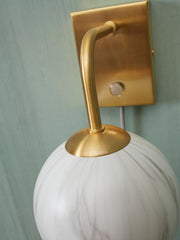 Detailansicht der Wandleuchte Carrara: goldener Messingarm mit weiß marmoriertem Glasschirm vor mintgrüner Wand.