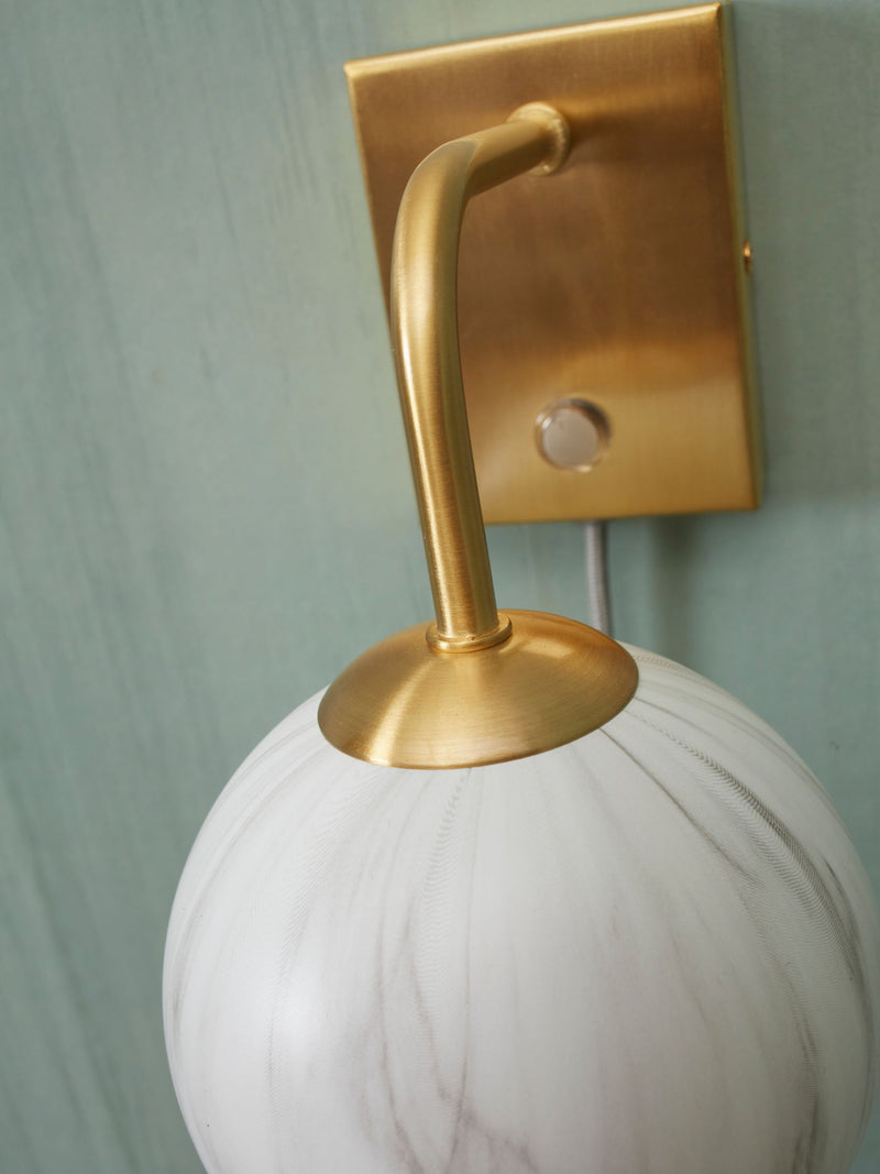 Detailansicht der Wandleuchte Carrara: goldener Messingarm mit weiß marmoriertem Glasschirm vor mintgrüner Wand.