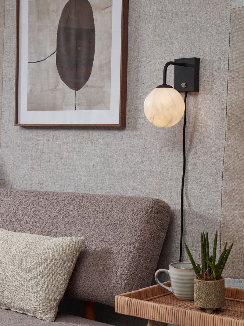 Wandleuchte It's about RoMi Carrara mit marmorierter Glaskugel und schwarzem Metallarm an einer Wand über einem Sofa.