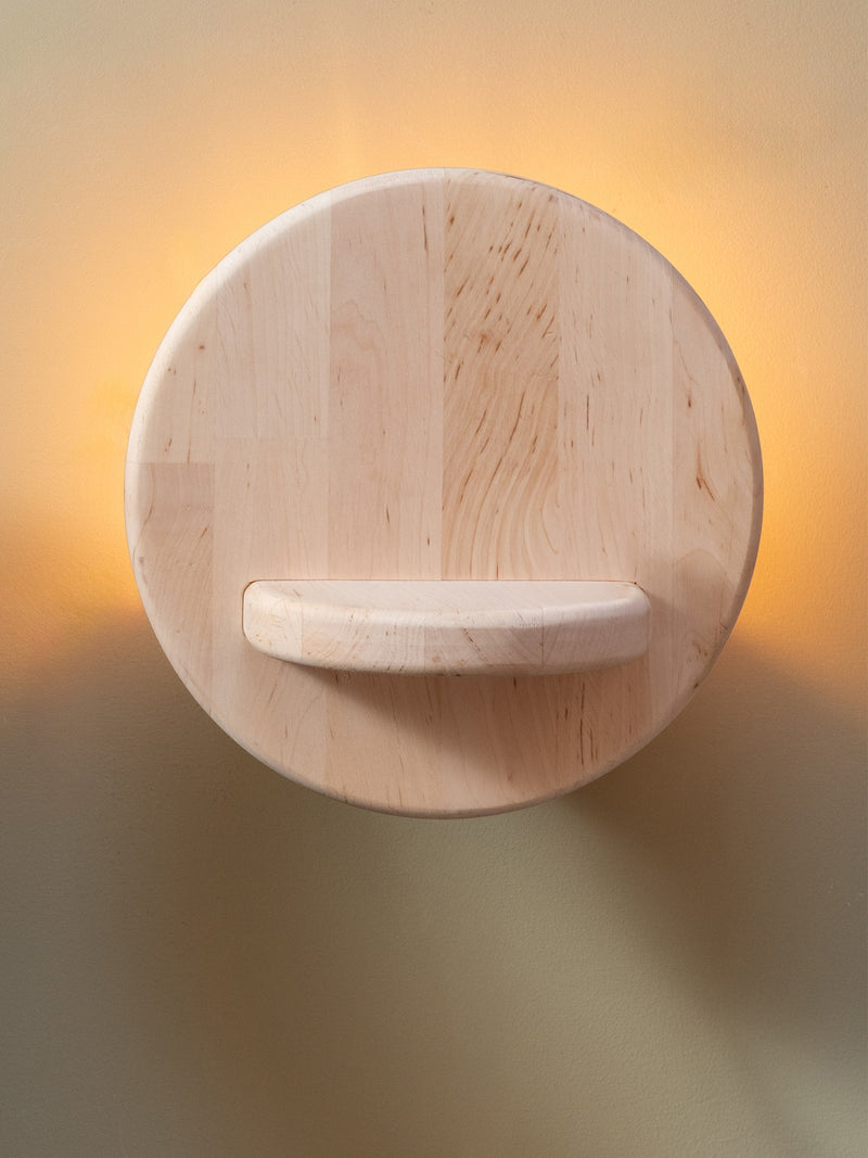 Runde Wandleuchte Corsica aus hellem Erlenholz mit angedeutetem Ablagesteg, sanft beleuchtet vor warmgelber Wand.