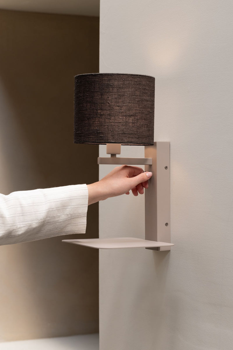 Die Wandleuchte RoMi Florence mit rundem, dunklem Leinenschirm und sandfarbenem Eisengestell mit kleiner Ablage, Hand am Schalter.