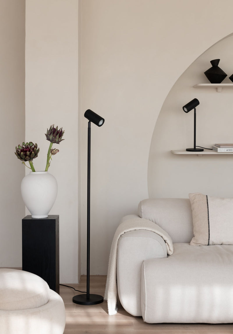 Schwarze Stehleuchte mit schmalem Stab und verstellbarem Spotkopf neben einem hellen Sofa und einer weißen Vase.