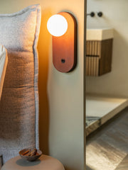 Wandleuchte Java von Good&Mojo mit ovalem Bambusgehäuse und weißem Glasschirm, im warmen Licht an der Wand neben einem Bett.