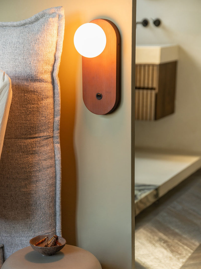 Wandleuchte Java von Good&Mojo mit ovalem Bambusgehäuse und weißem Glasschirm, im warmen Licht an der Wand neben einem Bett.