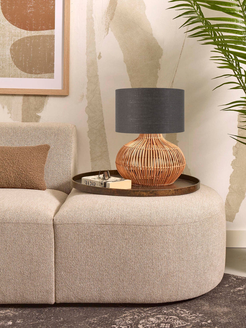 Tischleuchte Kalahari mit geflochtenem Rattan-Fuß und dunkelgrauem Leinenschirm, auf einem runden Tablett neben einem Sofa.