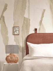 Tischleuchte Kalahari mit Rattan-Fuß und grauem Leinenschirm auf einem weißen Beistelltisch neben einem Bett.