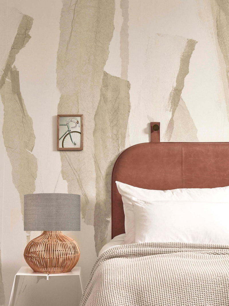 Tischleuchte Kalahari mit Rattan-Fuß und grauem Leinenschirm auf einem weißen Beistelltisch neben einem Bett.
