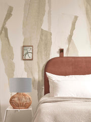 Good&Mojo Tischleuchte Kalahari aus Rattan mit grauem Leinenschirm auf weißem Beistelltisch neben einem Bett.