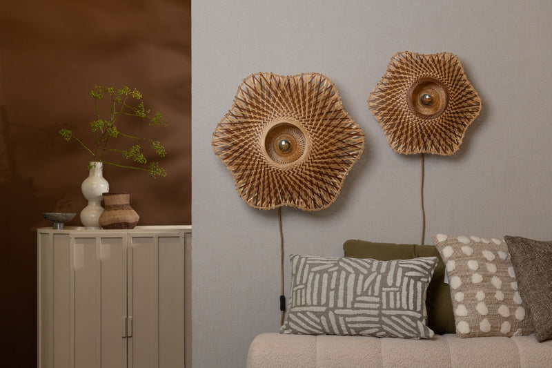 Zwei blütenförmige Wandleuchten Madura aus geflochtenem Bambus in Erdtönen hängen an einer hellen Wand über einem Sofa.
