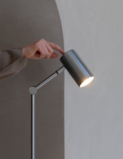 Eine Hand richtet den zylindrischen Schirm der Nickel-Stehleuchte RoMi Montreux aus, die Lampe leuchtet.