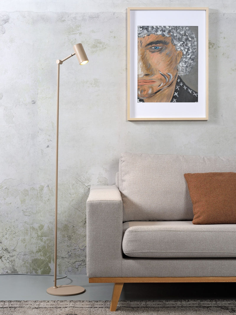 Stehleuchte Montreux in Sand mit verstellbarem Schirm neben einem Sofa, darüber ein gerahmtes Porträt an der Wand.