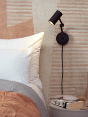 It’s about RoMi Wandleuchte Montreux aus Eisen in Schwarz, schwenkbar an der Wand über einem Bett mit Kissen.