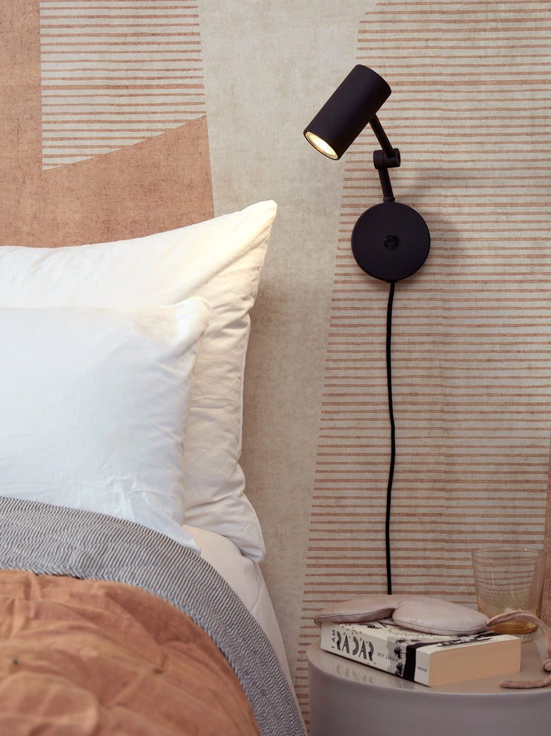 It’s about RoMi Wandleuchte Montreux aus Eisen in Schwarz, schwenkbar an der Wand über einem Bett mit Kissen.