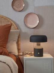 Die RoMi Tischleuchte Porto in Schwarz steht auf einem hellen Nachttisch neben einem Bett mit Rattan-Kopfteil, eingeschaltet.