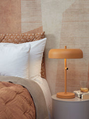 Senfgelbe Tischleuchte It's about RoMi Porto L mit rundem Schirm neben einem Bett mit gemustertem Kissen und Wandtapete.