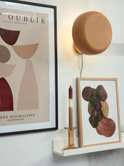 Die Wandleuchte „It's about RoMi Porto" in Senfgelb hängt rund über einem Wandbord mit Kunstdrucken und Kerze.