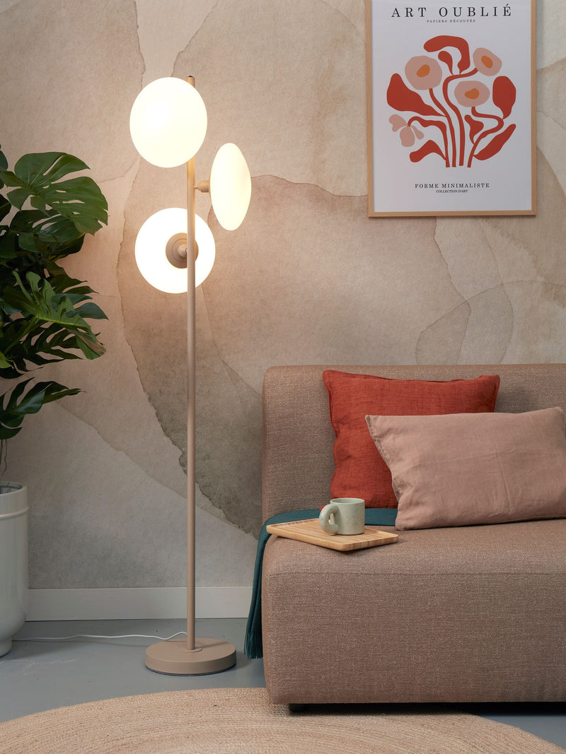 Stehleuchte Sapporo in Sand mit drei weißen Glaskugeln an einem schlanken Eisengestell, daneben ein Sofa.