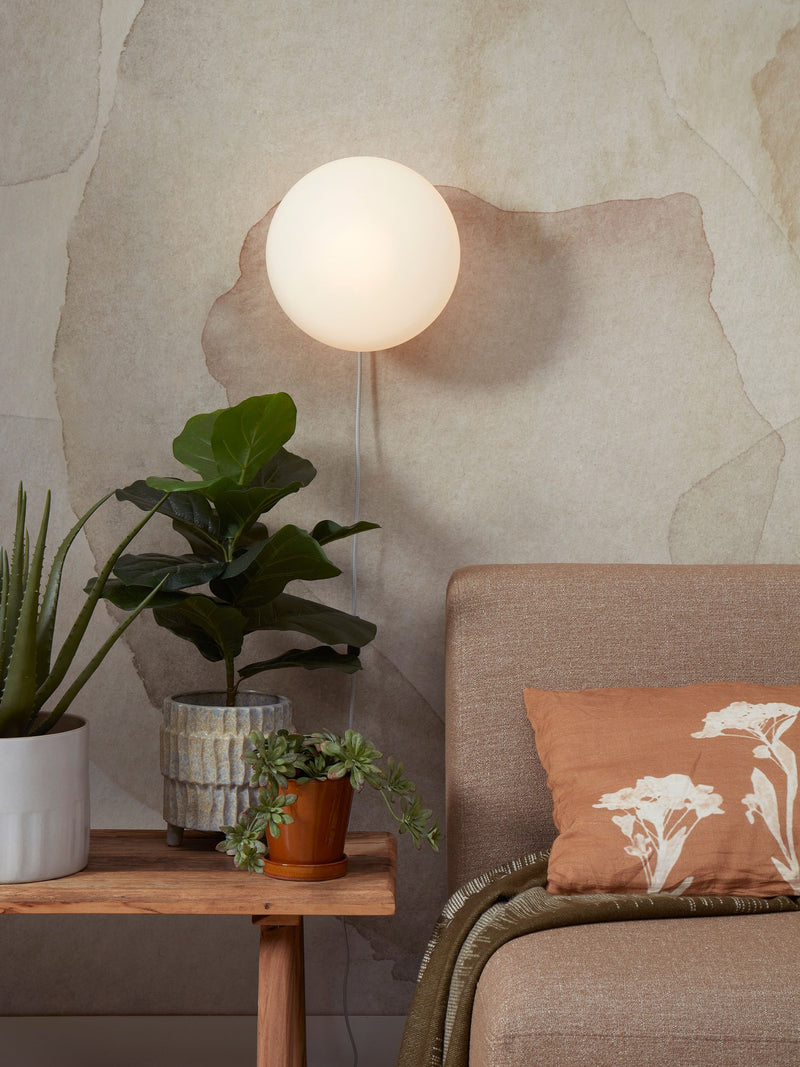 Wandleuchte RoMi Sapporo aus Glas in Weiß/Sand: runde, weiß leuchtende Glaskugel an der Wand, daneben Pflanzen und Sofa.