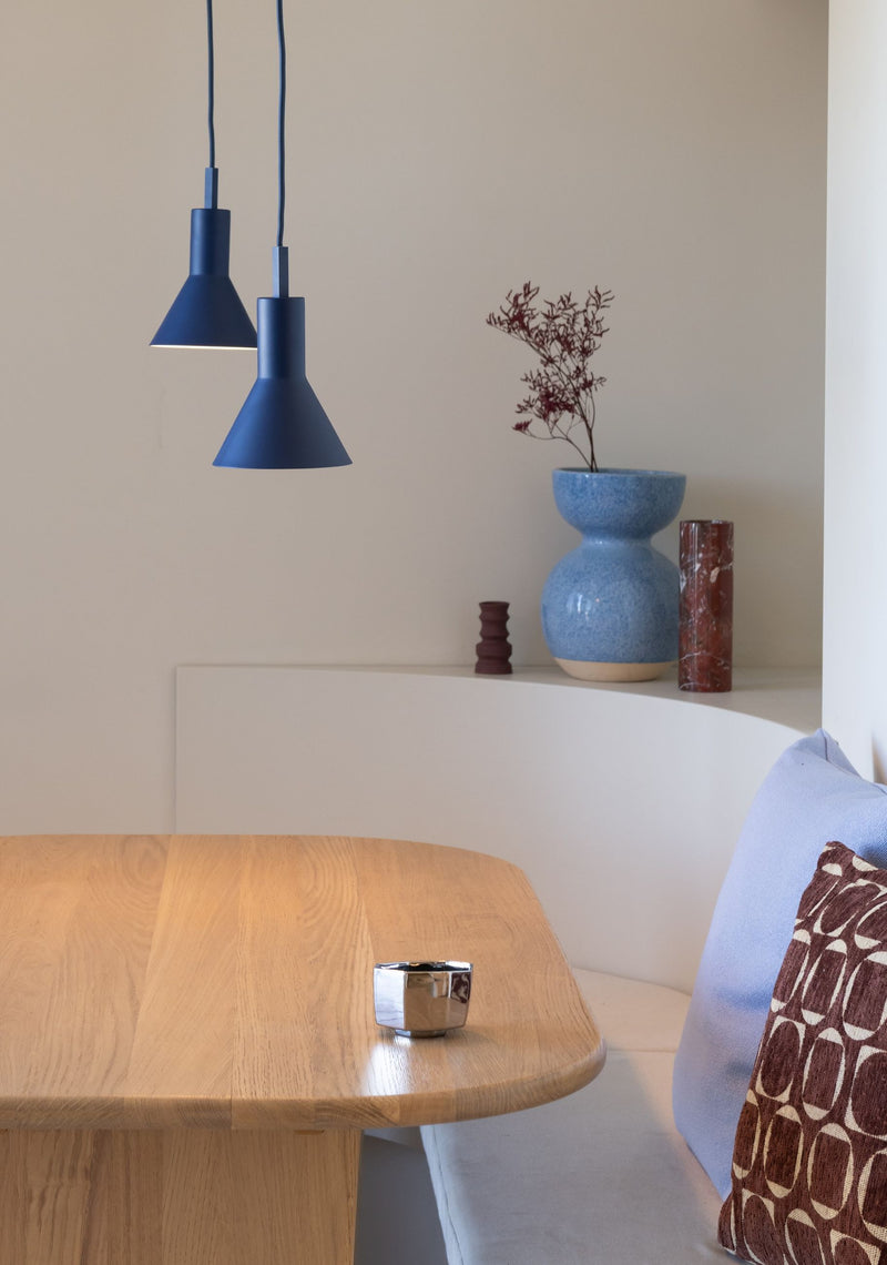 Zwei dunkelblaue Hängeleuchten „It's about RoMi Stavanger“ aus Metall mit konischen Schirmen über einem Holztisch mit Bank.