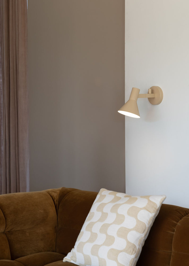Die RoMi Wandleuchte Stavanger in Beige mit schwenkbarem Schirm hängt an einer Wand neben einem braunen Samtsofa.