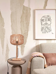 Tischleuchte Tanami von Good&Mojo mit geflochtenem Rattan-Schirm und Bambusfuß auf einem Beistelltisch vor einer beigen Wand.