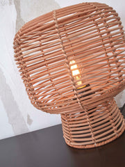 Good&Mojo Tischleuchte Tanami aus Rattan in Natur: runder Rattanschirm mit leuchtender Glühbirne auf dunklem Sockel vor heller Wand.